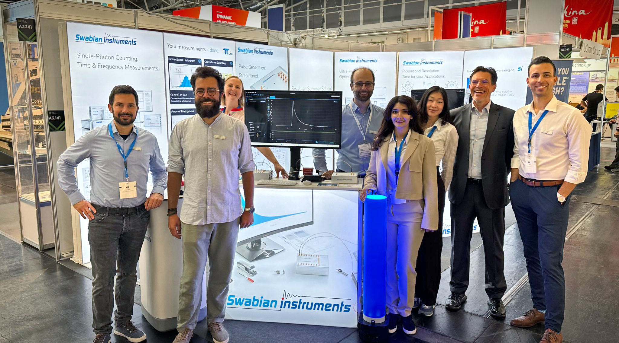 The Swabian Instruments team (eight members) in front of the Swabian Instruments exhibition booth, which shows banners detailing the device specs and a live demonstration, at the Laser World Photonics 2025. The live demonstration shows a Fluorescence Lifetime experimental setup, in which a pulsed laser sends a pulse into a sample, and a SPAD detects the arrival of the emitted photon. The Time Tagger Lab is shown on a monitor, with the histogrammed curve of the fluorescence lifetime of the sample.