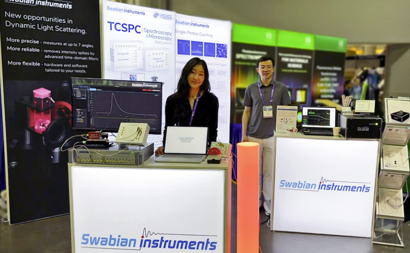 Conference booth display for Swabian Instruments featuring Ginger (Tingting) Geng on the left and Yizhou Wang on the right, standing beside illuminated counters, monitors, and photonics instrumentation, with signage for dynamic light scattering and TCSPC in the background.