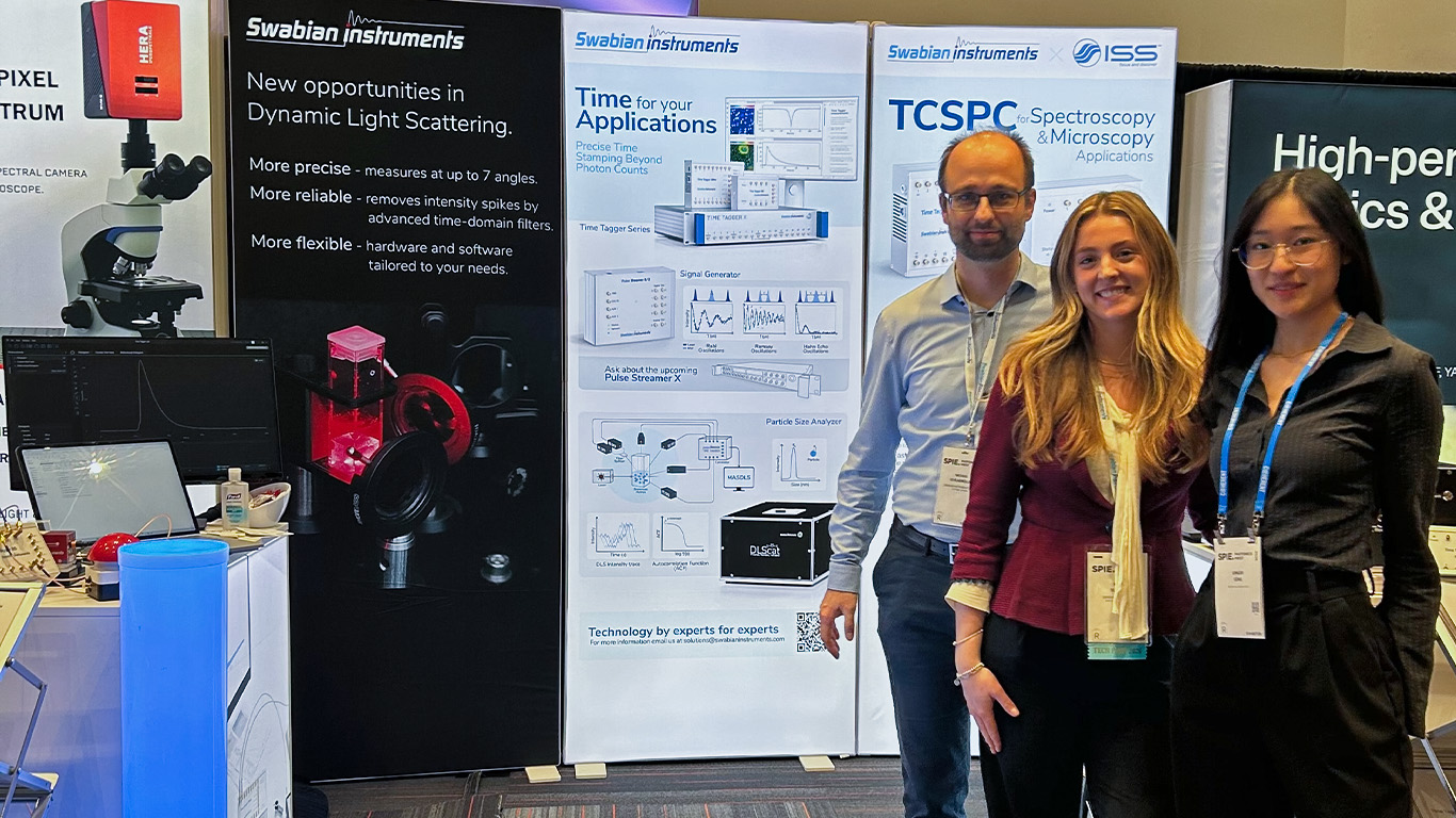 Swabian Instruments team members at one of the exhibition booths for BiOS, standing in front of the informational walls about TCSPC and Dynamic Light Scattering using the Time Tagger Series. From left to right: Michael Schlagmüller, Maré Sutphen, and Ginger Geng.