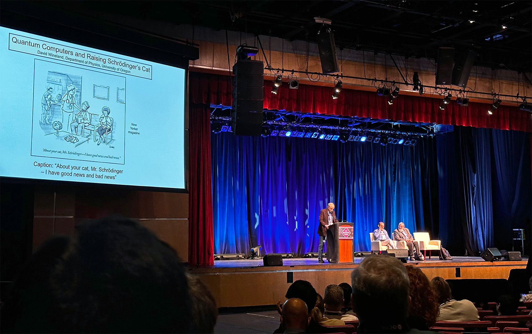David J. Wineland stands at a podium on the Kiva Auditorium stage during IEEE Quantum Week 2025. A large slide titled “Quantum Computers and Raising Schrödinger’s Cat” shows a New Yorker-style cartoon with the caption “About your cat, Mr. Schrödinger—I have good news and bad news.” Two fellow panelists (one being Philips) sit on white chairs to the right; the audience is visible in the foreground.