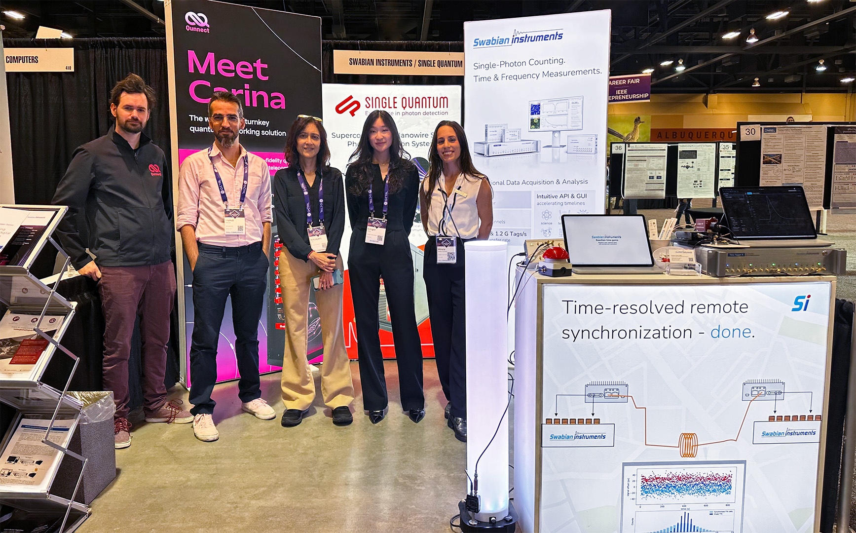 Swabian Instruments, Qunnect, and Single Quantum team members (five total) stand together at a joint booth in the IEEE Quantum Week 2025 exhibition hall in Albuquerque. Behind them, stand three backdrops. Qunnect’s backdrop to the left reads “Meet Carina,” Single Quantum’s backdrop in the middle displays the company name and logo, and Swabian Instruments’ backdrop to the left reads “Single-Photon Counting Time & Frequency Measurements.” In the foreground, Swabian Instruments’ front counter reads “Time-resolved remote synchronization — done.” and shows a basic diagram of a remote sync setup with a laptop illustration between two White Rabbit nodes and Time Taggers connected by optical fiber. Overhead aisle signs and poster boards are visible in the convention hall.