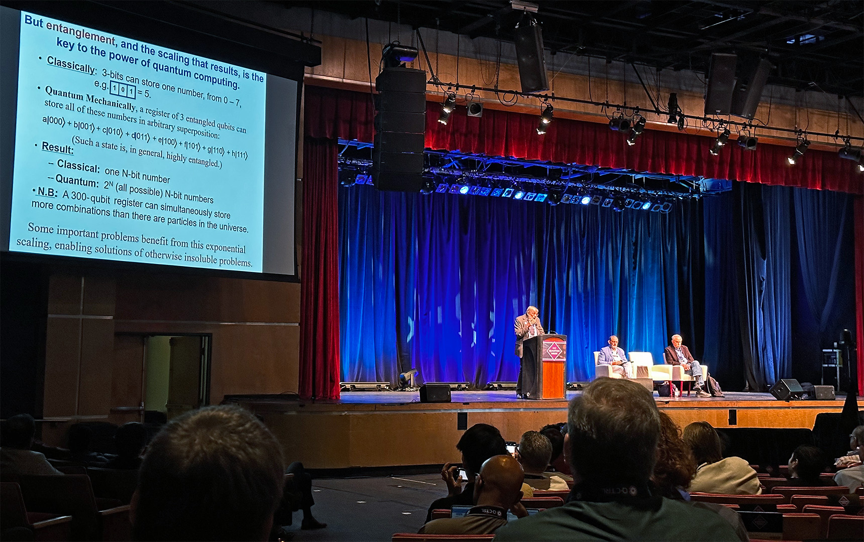 William D. Phillips presents from a podium on the Kiva Auditorium stage at IEEE Quantum Week 2025. A projected slide reads “But entanglement, and the scaling that results, is the key to the power of quantum computing,” contrasting classical vs. quantum register capacity and noting exponential scaling. Two fellow panelists (one being Wineland) sit on white chairs to the right; the audience is visible in the foreground.