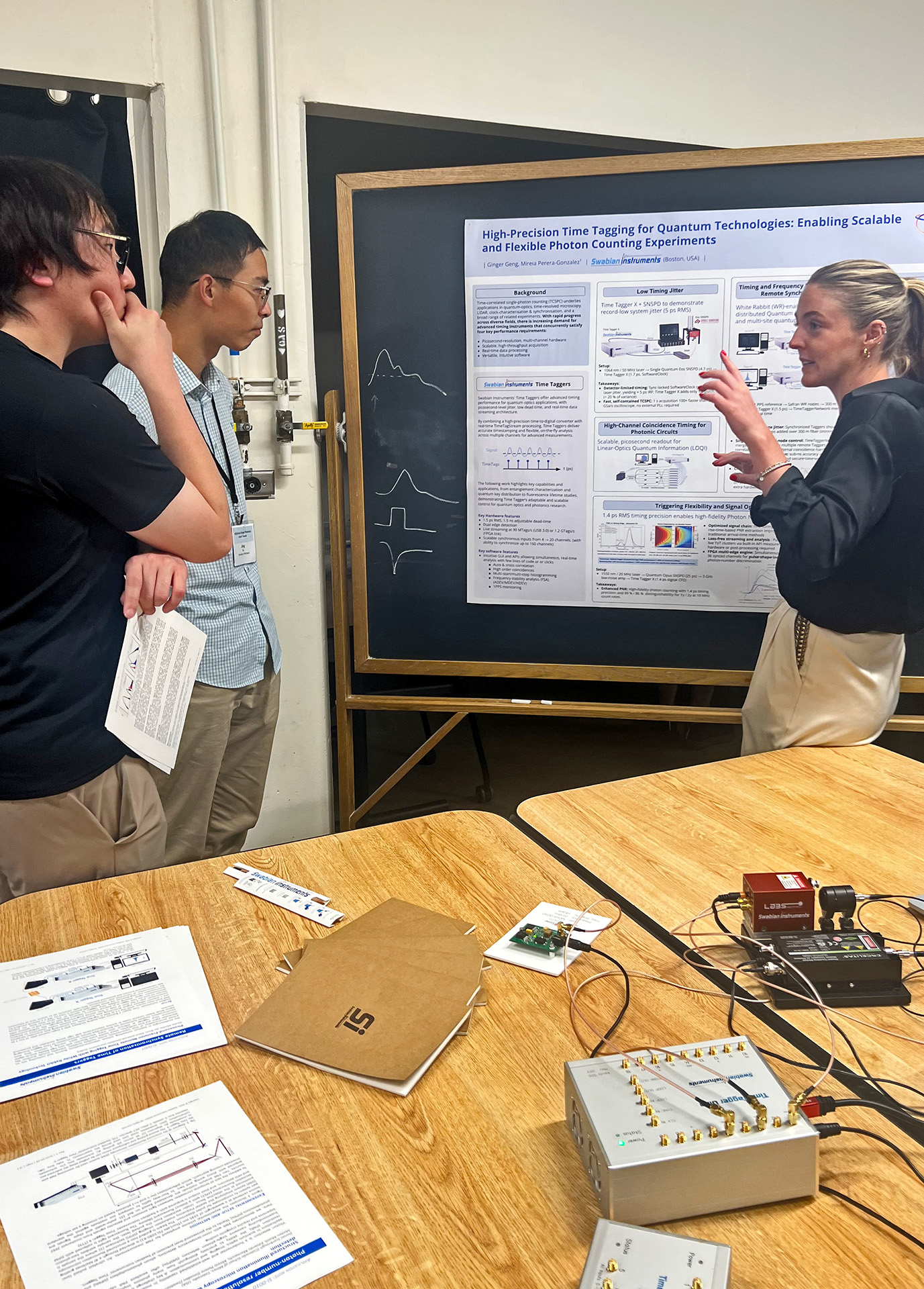 Swabian Instruments personnel using a poster to explain the precision and capabilities of the Time Taggers to two Short Course participants. The table at which they are standing shows a Fluorescence Lifetime demonstration and a Frequency Stability Analysis demonstration using the Time Tagger Ultra and Pulse Streamer.