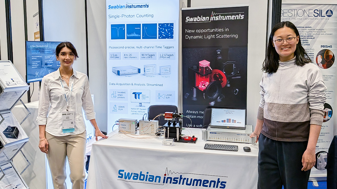 Swabian Instruments team members standing at their exhibition booth at SPIE Photonics Europe in Strasbourg, showcasing single-photon counting and time tagging solutions with a live demo setup and measurement equipment.